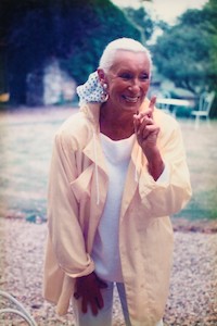 Mita wearing a pastel yellow rain jacker, white blouse and jeans, looking off camera and pointing her hand as a fun gesture whilst smiling
