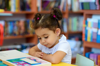 young girl wearing a white t-shirt with her hair in bunches, reading a book in the library