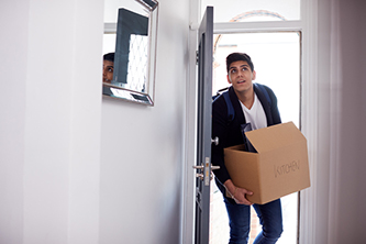 Young man with dark hair wearing a check shirt, holding a cardboard box whilst carrying through his new home