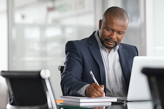 Man in front of laptop wearing a formal suit and writing on a notepad