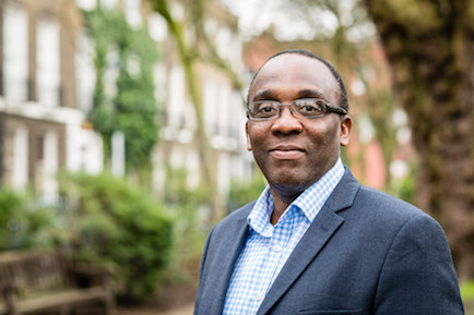 Demi Lawal, wearing glasses and a suit and standind in front of houses