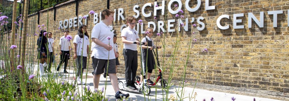 School children walking and using scooters as they walk past a wall with the words 'Robert Blair School and Children's Centre' on it 
