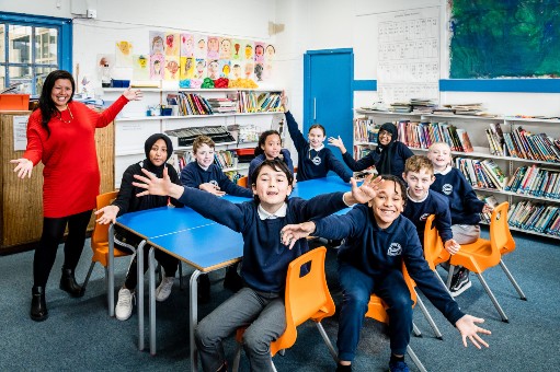 A classroom of smilingchildren sitting around a table arms outstretched to the camera with their teacher standing with the same welcome gesture